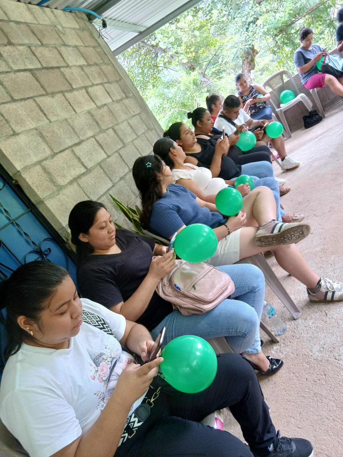 Women sitting in a row on a covered porch in San Salvador, each holding a green balloon