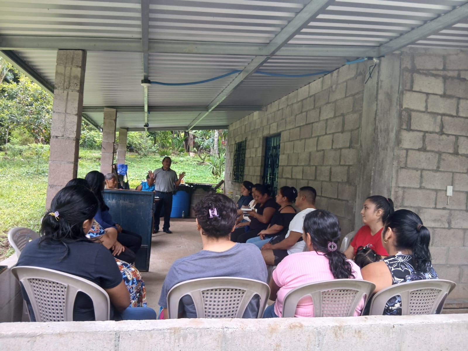 A facilitator speaking to a group seated in plastic chairs on an open porch in San Salvador